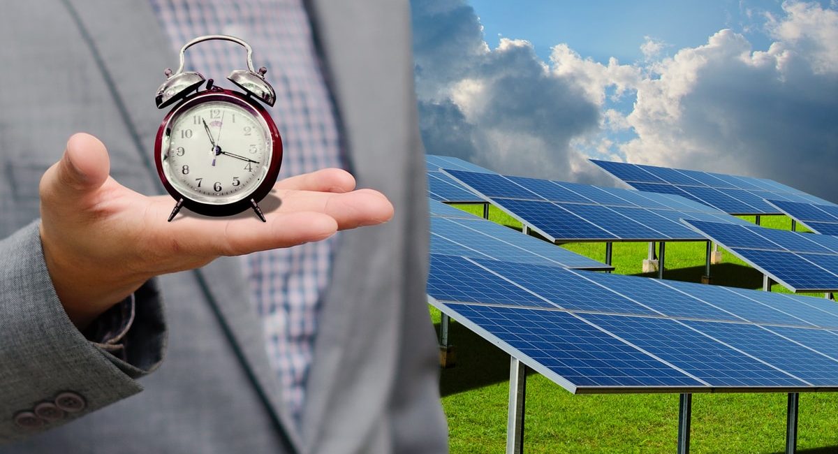 un uomo tiene un orologio in mano con lo sfondo un impianto fotovoltaico. Illustra che per avere un impianto attivo bisogna attendere i tempi di connessione e autorizzazione.